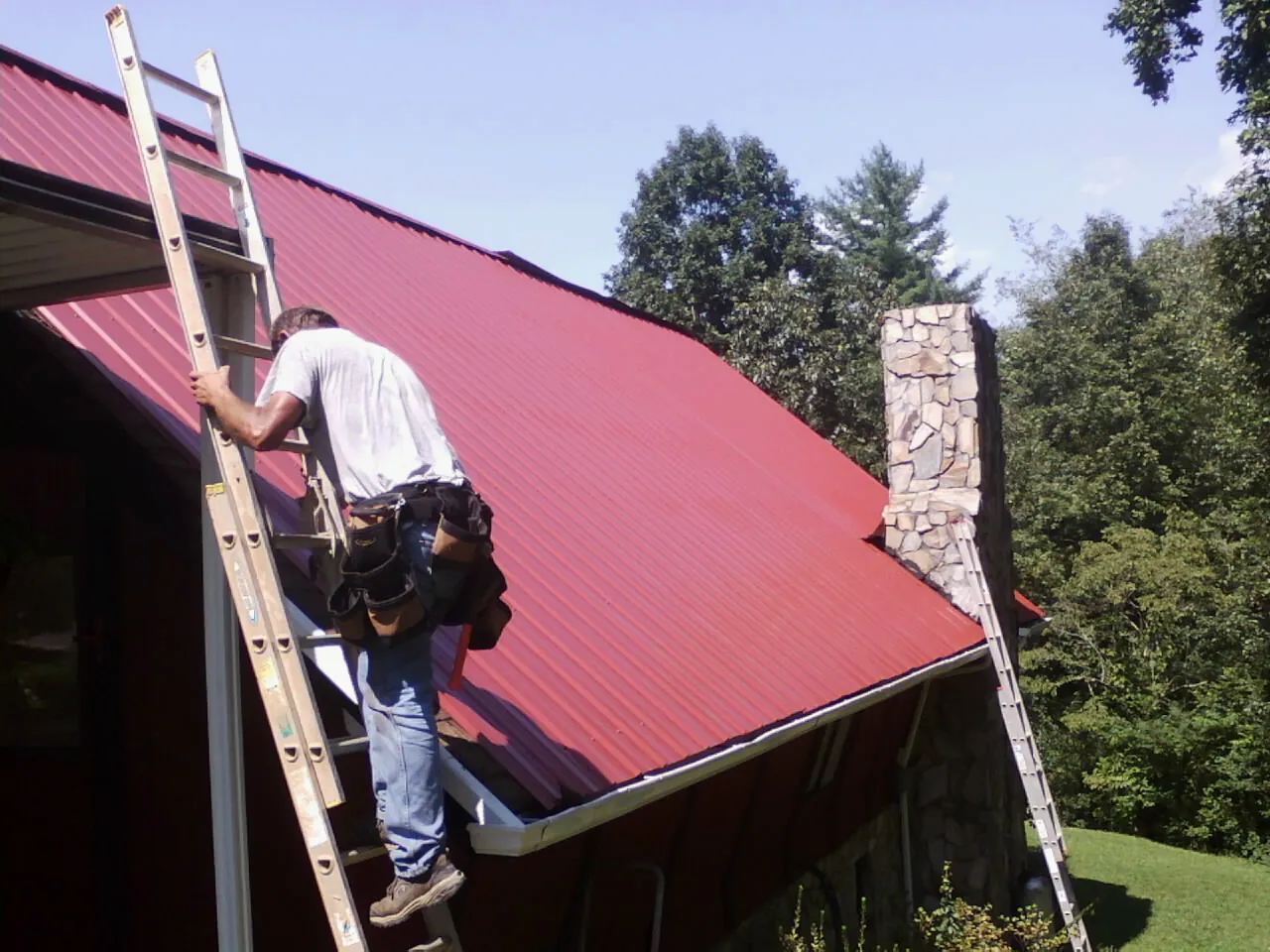 Roofer on a ladder beside a red metal roof and stone chimney — Soffit & Fascia Repair by Roofing Brothers in Marseilles